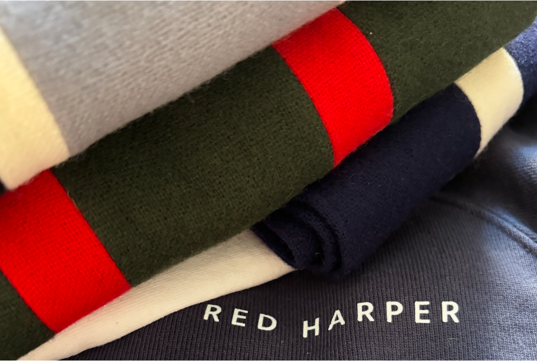 Close-up of folded clothing in textured fabric. A dark blue garment with the printed text “RED HARPER” is layered beneath a cream-colored piece and a dark green fabric with a bold red stripe.