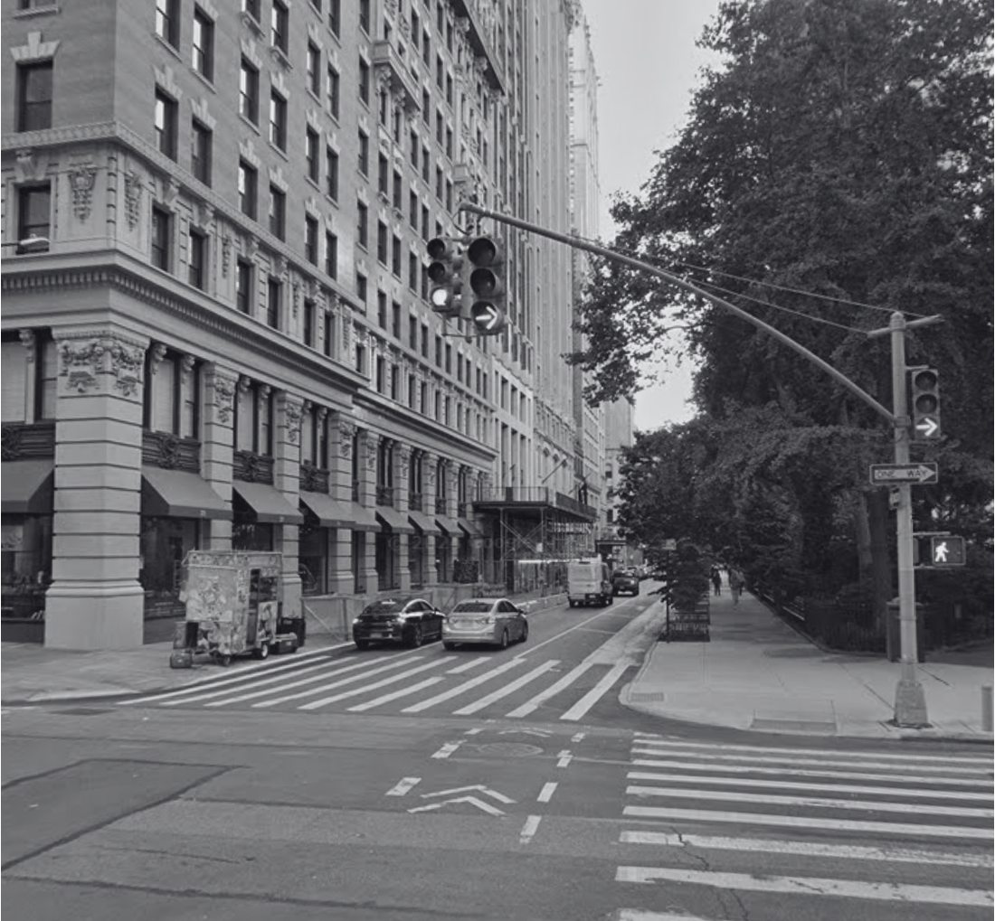 Black-and-white street view of a Manhattan intersection with tall historic buildings, a traffic light overhead, crosswalk markings on the asphalt, and a tree-lined sidewalk beside a city park.